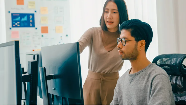 A colleague leaning in to help someone working at a desktop workstation